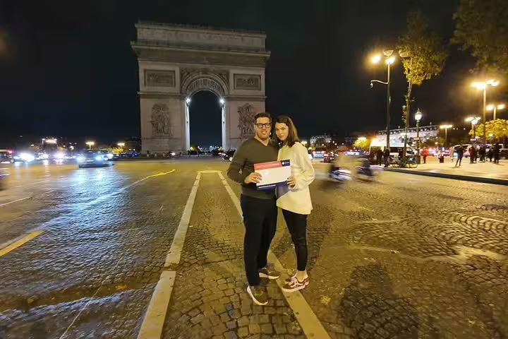Couple posing by Arc de Triomphe at night on private Paris sightseeing tour with city lights and traffic