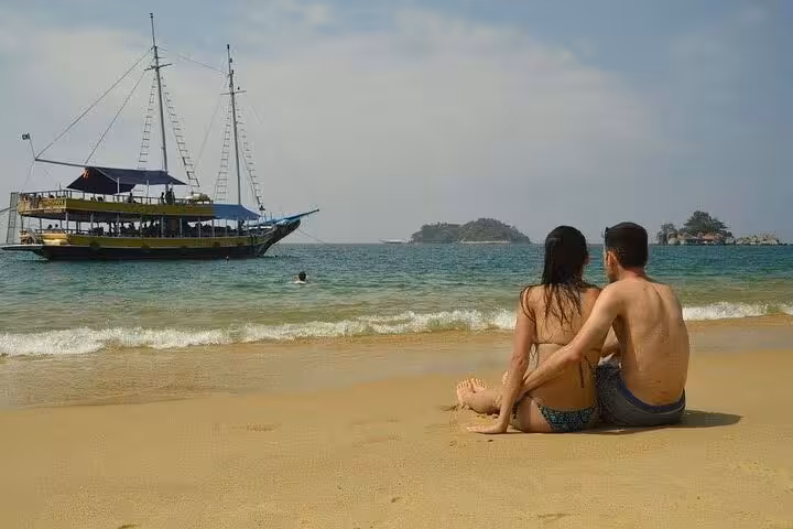 Couple relaxing on a sandy beach with a scenic view of a sailboat at sea, part of the Paraty tours experience.