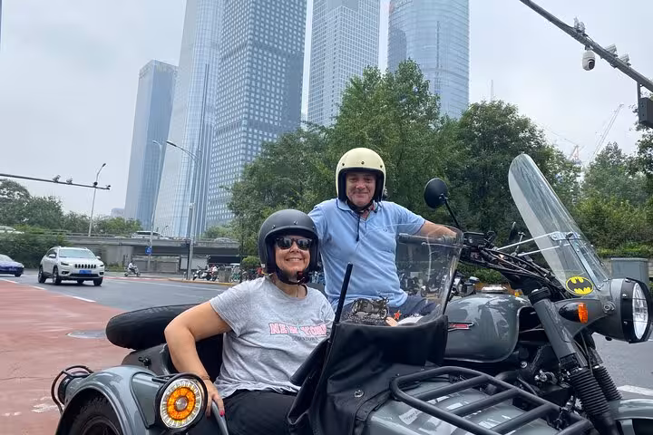 Smiling couple on a Beijing sidecar tour, set against the backdrop of modern skyscrapers in the cityscape.