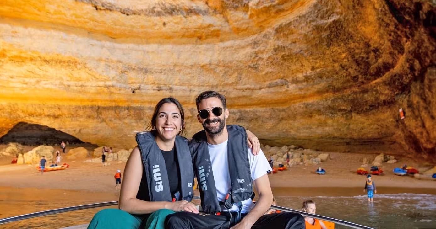 Smiling couple on a private Benagil cave boat tour, wearing life jackets with golden Algarve sea cave beach behind