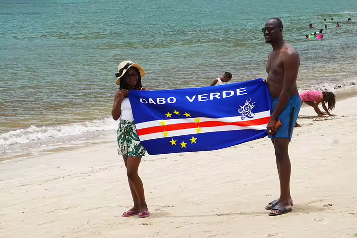 Couple holding the Cabo Verde flag on the sunny beach of Boa Vista, with turquoise waters in the background.