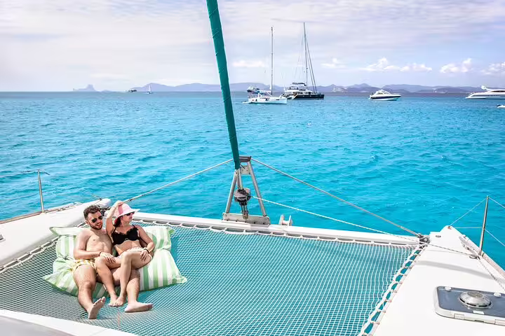 Couple relaxing on a catamaran net with turquoise waters and distant sailboats, enjoying a small group trip around Formentera and Ibiza.