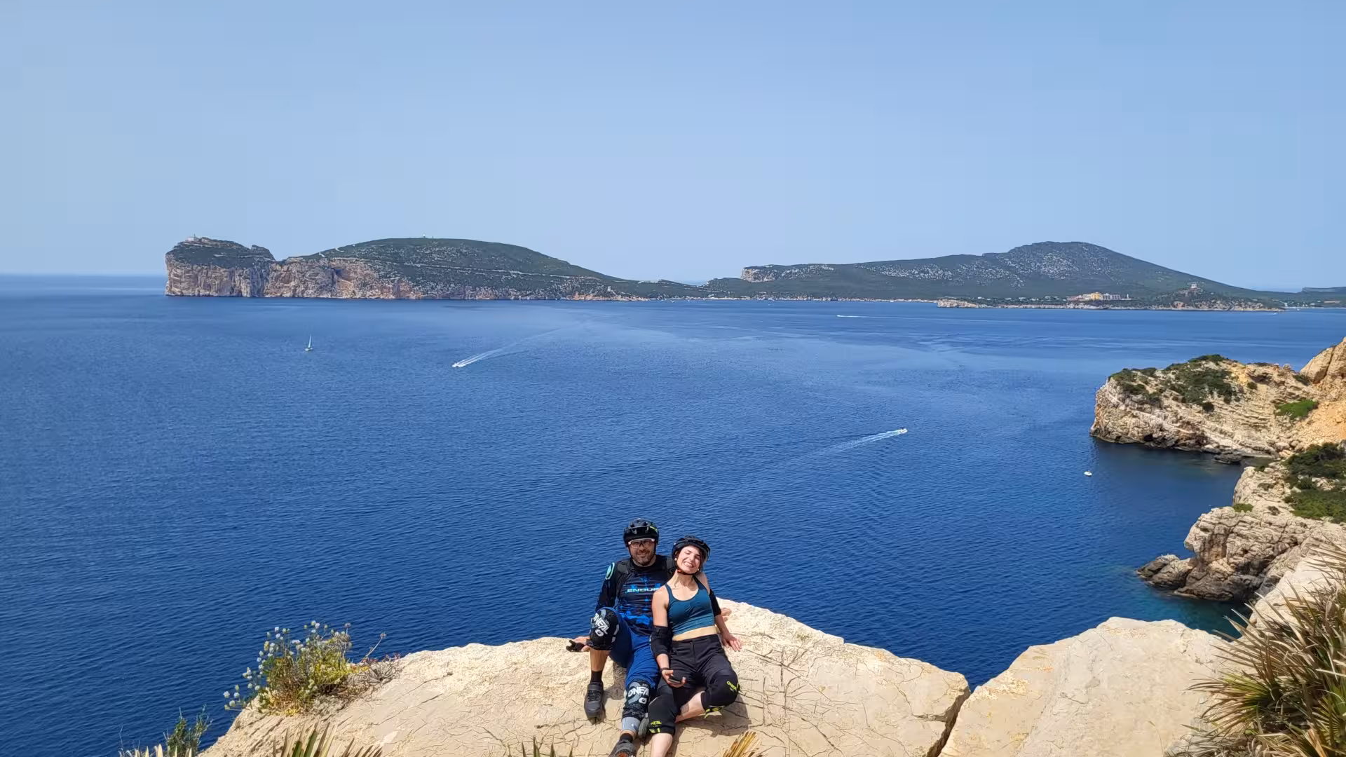 Couple enjoying a scenic view from a cliff during a guided e-bike tour along the Alghero coastline.
