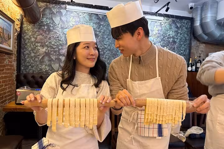 Couple in Florence pasta making class holding fresh homemade pasta with a chef in a cozy kitchen setting.
