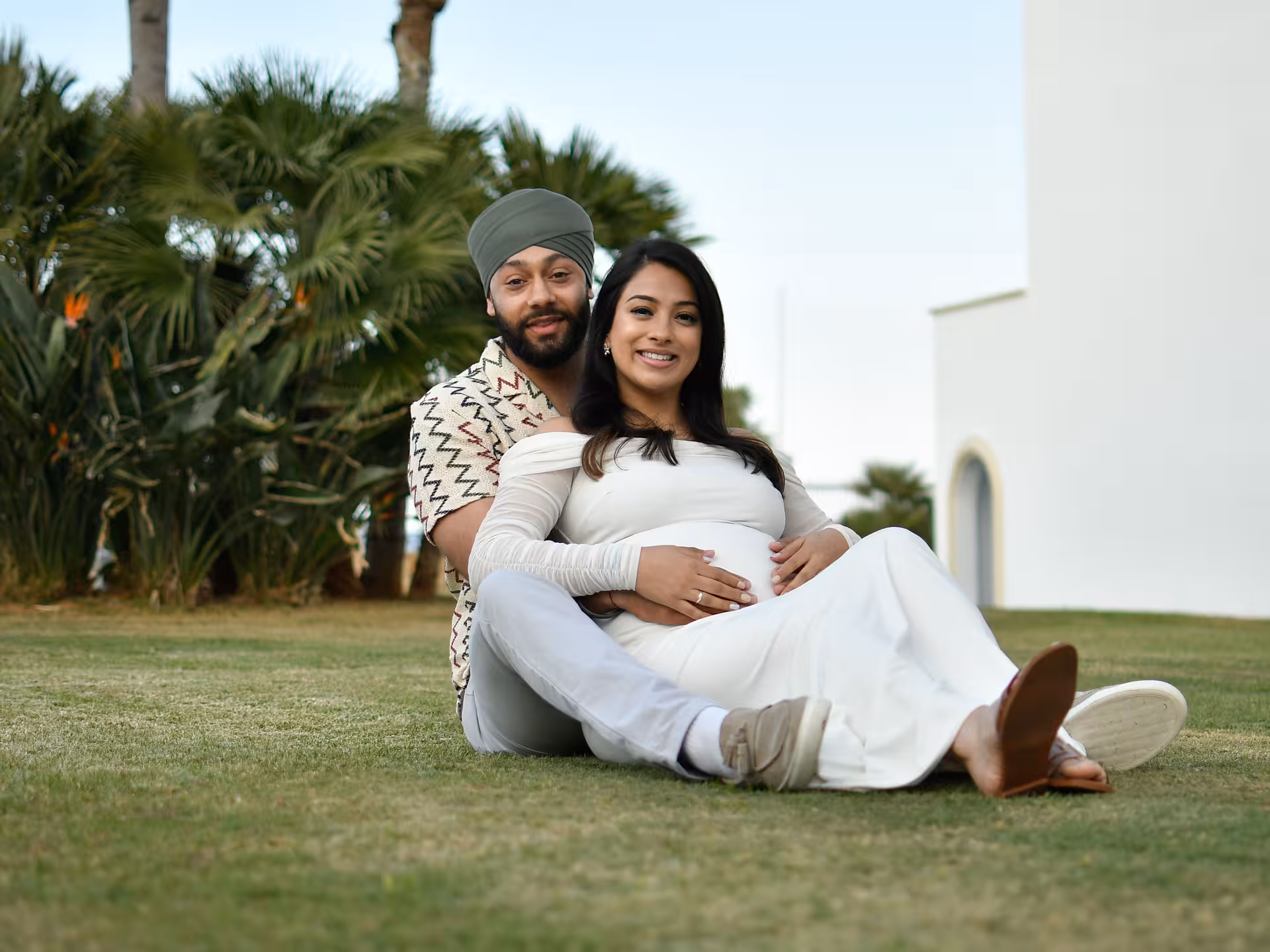 Couple sitting on grass in Heraklion garden, woman cradling belly, capturing intimate moment in private photo session.