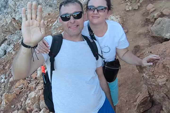 Couple hiking on rocky trail at Seitan Limania with backpacks, enjoying a sunny day at the beach.