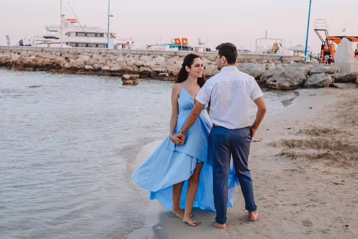 Couple holding hands strolls along Ayia Napa beach with boats in the background during a private photoshoot.