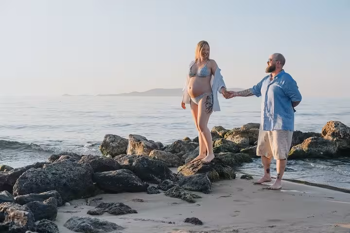 Couple stands on rocky Gouves shore, enjoying a maternity photoshoot with a scenic sea backdrop.