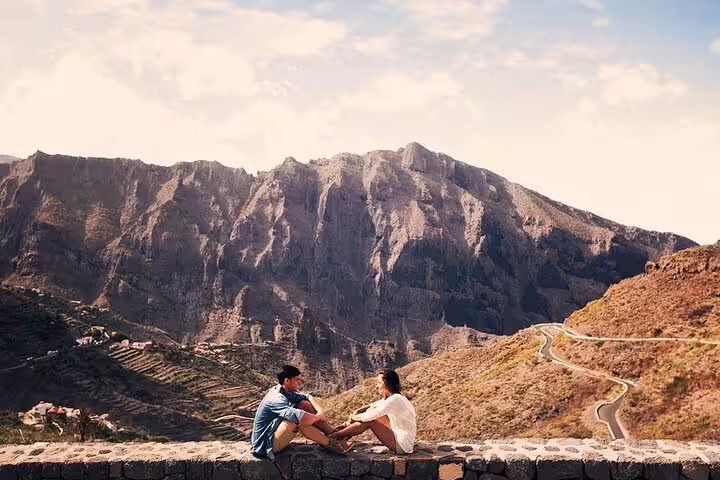 Couple sitting on a stone wall, embracing the breathtaking mountain views of Garachico, Tenerife on a sunny day.