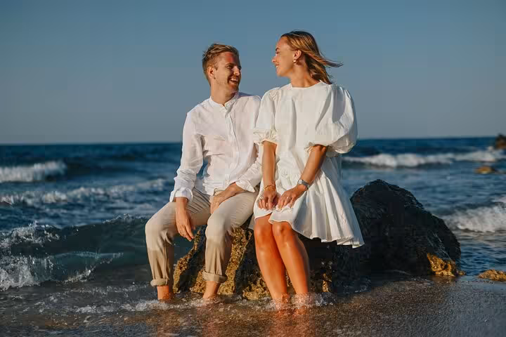 Smiling couple sitting on rocks with ocean waves at Agios Nikolaos beach during an exclusive photoshoot session.