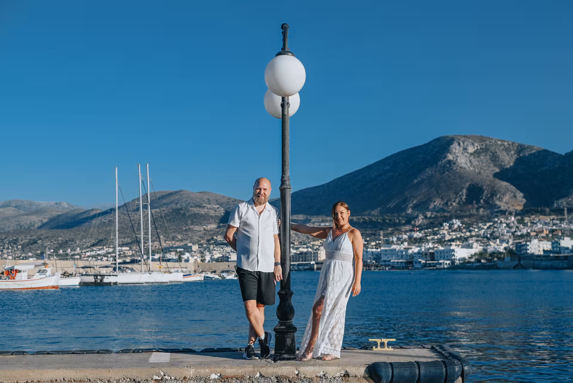 Couple posing by a lamppost with scenic views of the Port of Hersonissos and mountains in the background.