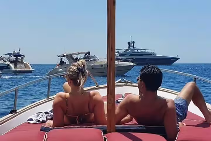 Couple sunbathing on a private Capri boat tour, passing luxury yachts on clear blue waters near the island