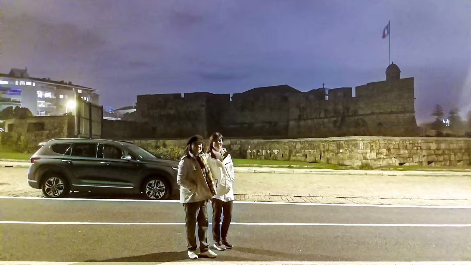 Couple enjoying a private night tour in Porto, standing near historic Castelo do Queijo, illuminated under a twilight sky.