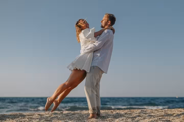 Couple embracing joyfully on sandy Agios Nikolaos beach during private photoshoot, clear blue sky and sea backdrop.