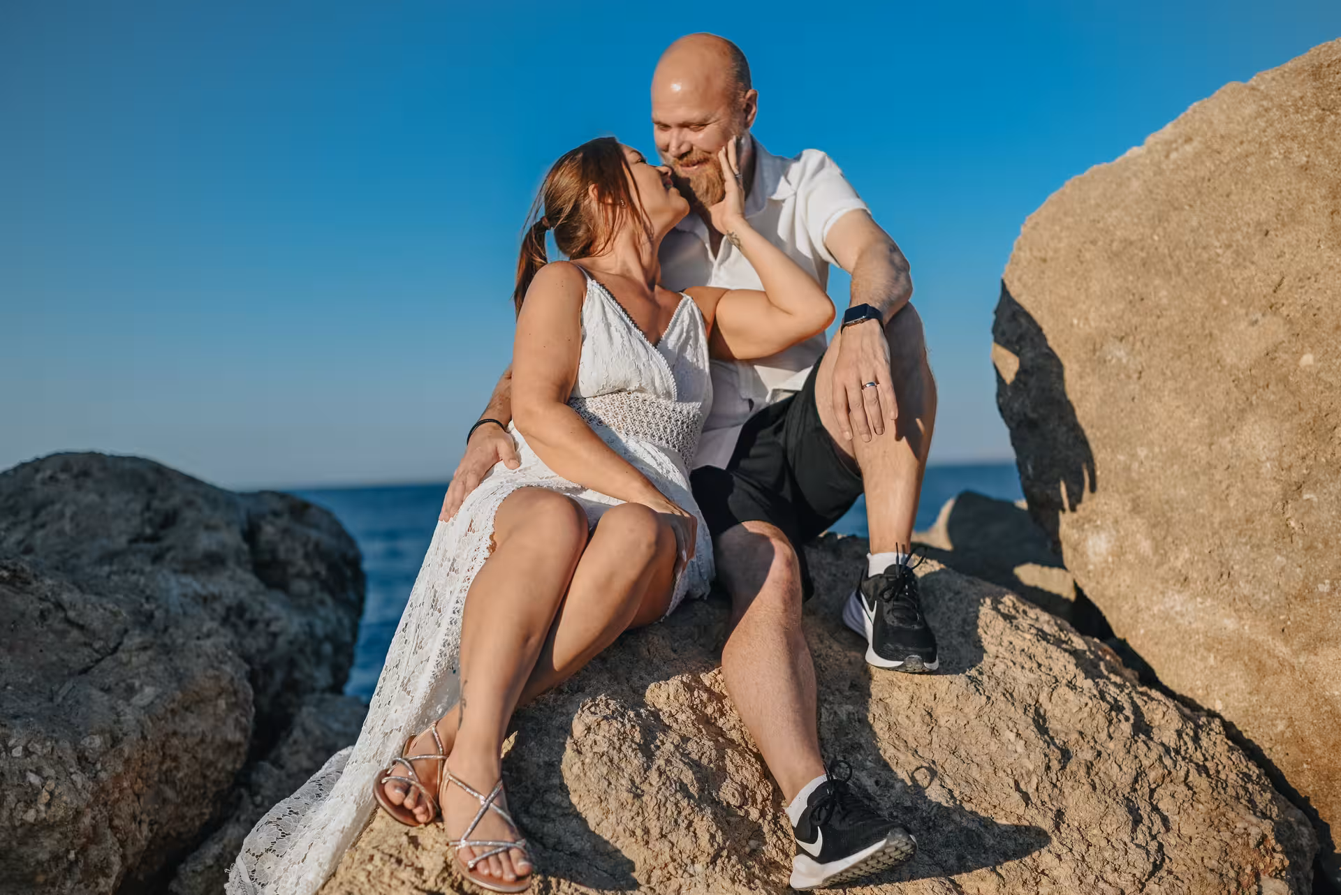 A couple shares a romantic moment on sunlit rocks by the sea at Hersonissos Port, perfect for a private photoshoot.