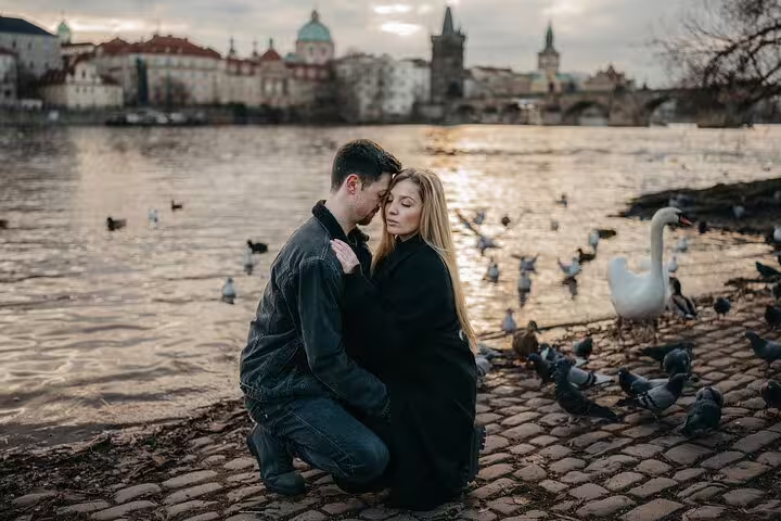 Couple embraces by the Vltava River with swans and Prague skyline, capturing a romantic photoshoot moment.