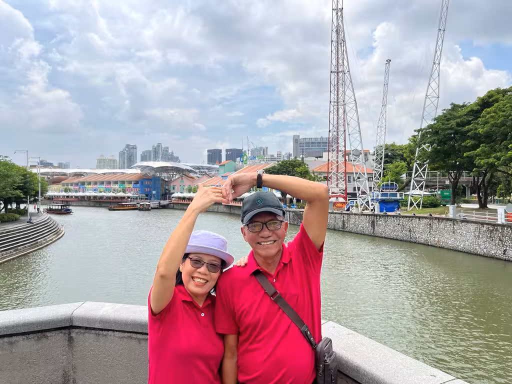 Couple makes heart shape with hands on Singapore River bridge, part of a romantic city tour.
