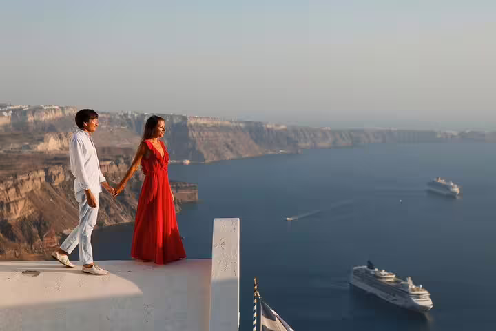Couple in elegant attire holding hands on a Santorini rooftop with stunning caldera and cruise ship views.