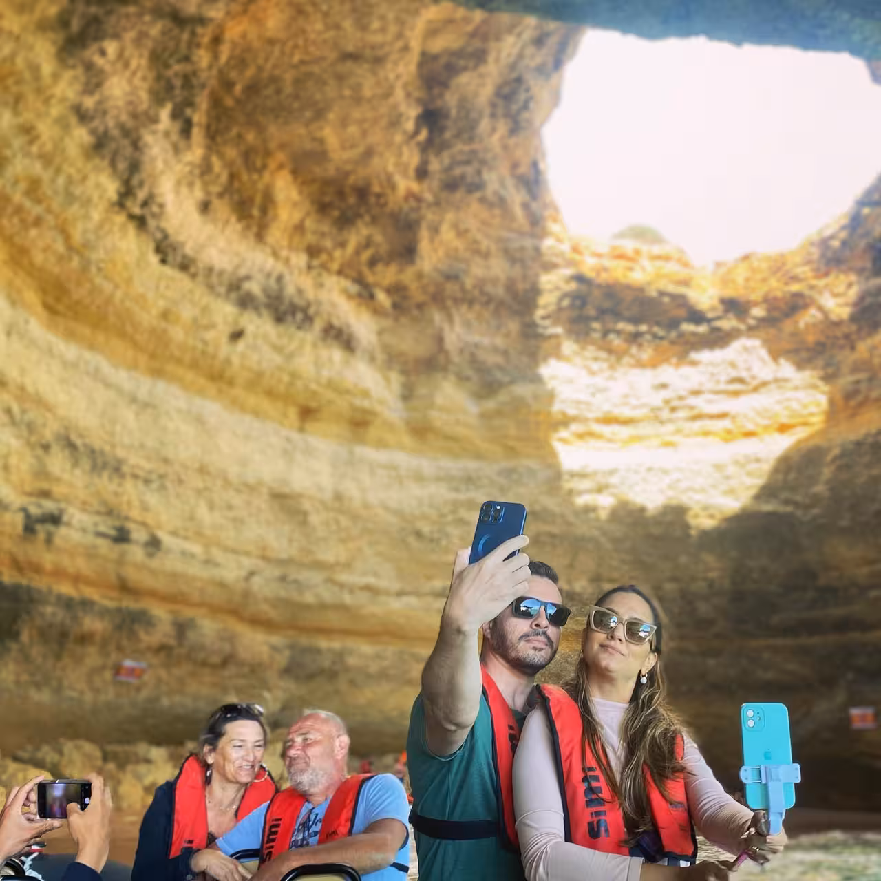 Couple taking selfies inside Benagil Cave on a private Algarve boat tour, wearing life jackets beneath the sunlit rock dome