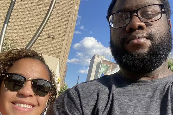 Two people taking a selfie outdoors in Philadelphia with city buildings and blue sky behind them.