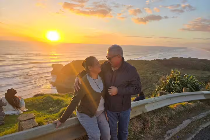Happy couple embracing at sunset on Auckland's scenic coastline, highlighting a romantic tour with winery and thermal pool.