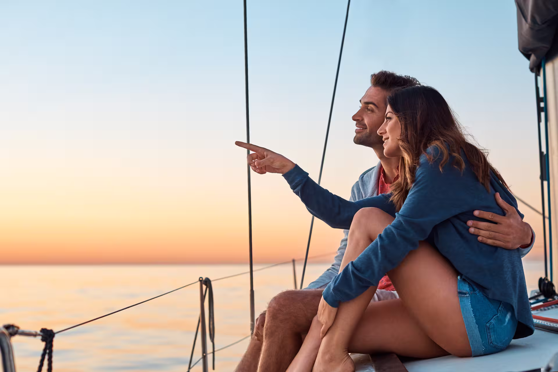 Couple enjoys a serene sunset sailboat experience, pointing at the horizon, perfect for a romantic 2-hour tour with swim.