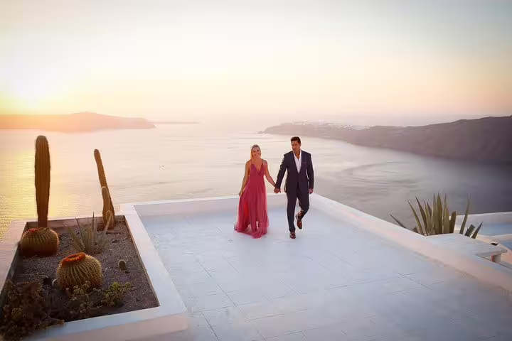 Couple in elegant attire walking hand in hand on a Santorini terrace at sunset, overlooking the Aegean Sea.