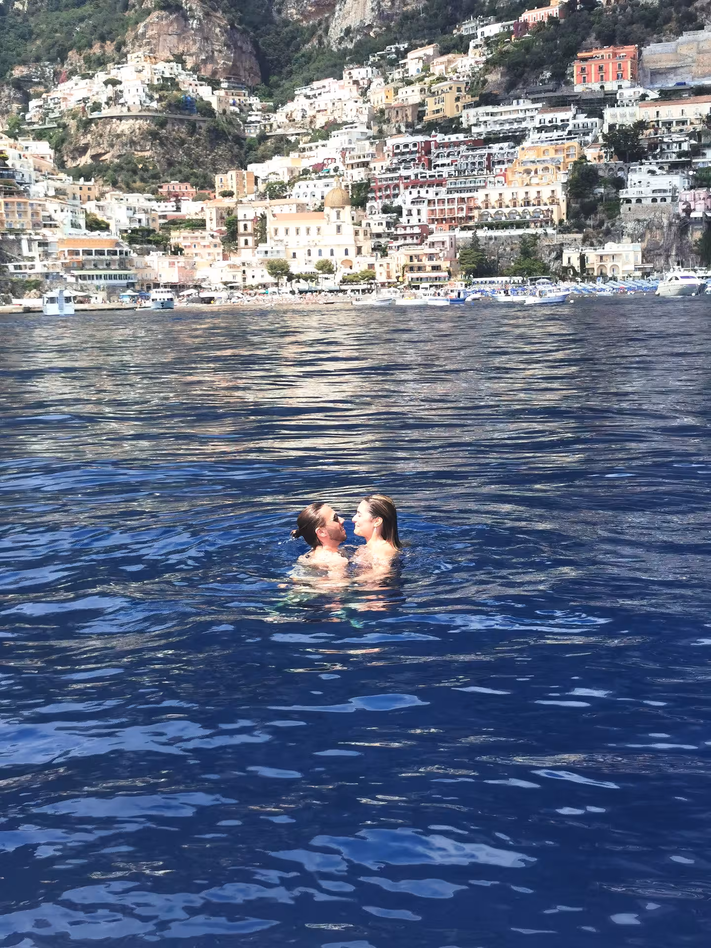Couple swimming in clear blue waters with Positano's colorful cliffside buildings in the background.