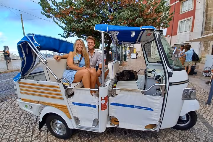 Couple seated in tuk-tuk for Lisbon 2-hour sightseeing tour, riding along riverside streets near Tagus River