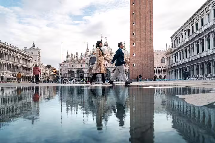 Couple holding hands and walking through St. Mark's Square in Venice, reflecting in puddle with historic architecture.