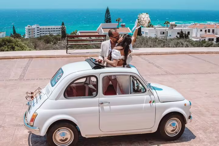 Couple celebrating in a vintage car with a bouquet, overlooking Ayia Napa's stunning coastline.