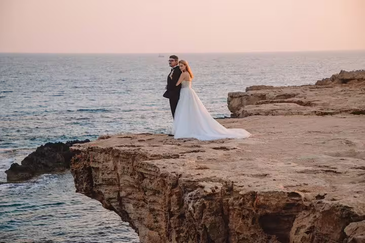 Couple in wedding attire embracing on a dramatic cliffside at sunset in Ayia Napa for a private photoshoot.