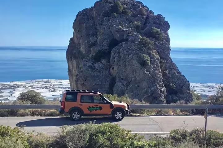 Adventure Tours vehicle driving along a scenic coastal road near a massive rock formation in Crete, Greece.