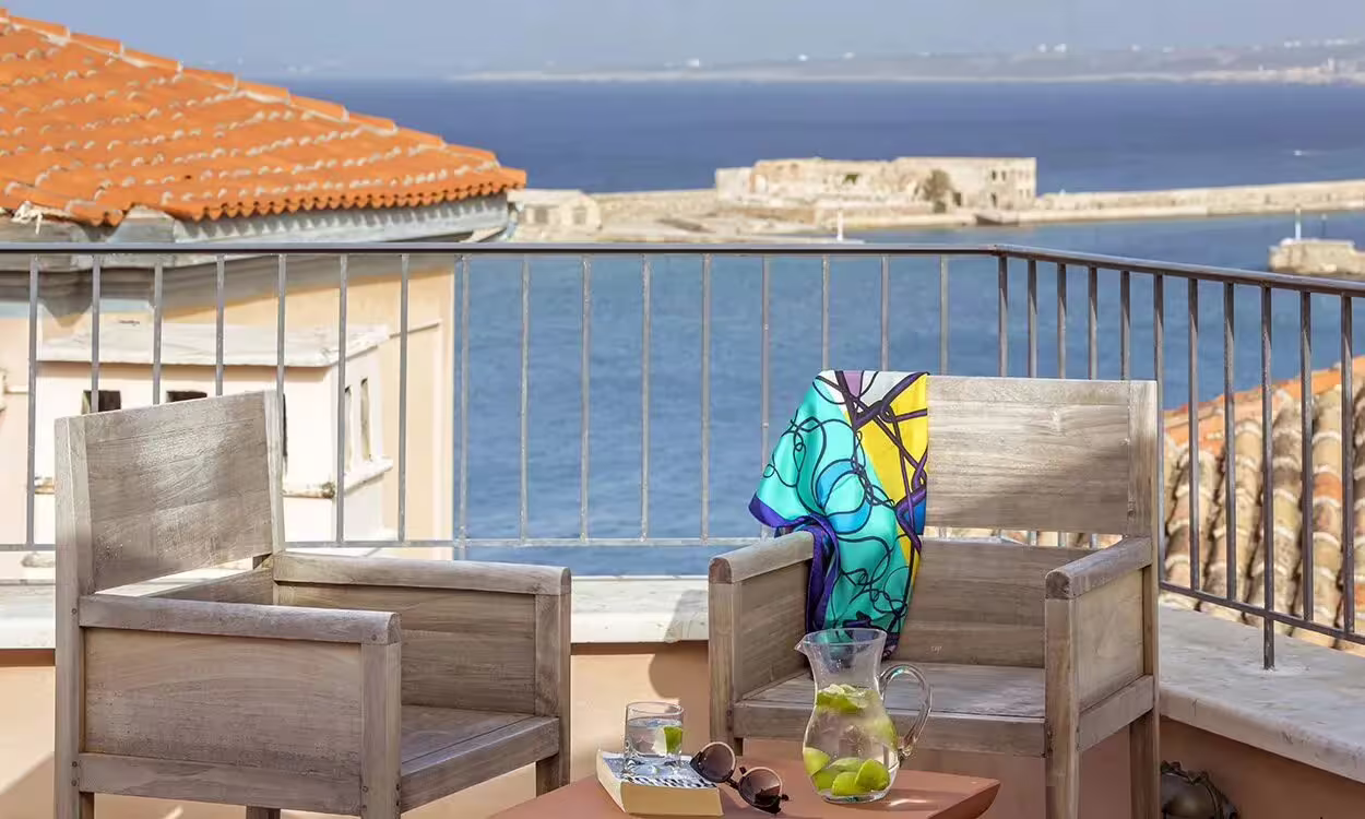 Charming terrace with sea views in Chania, featuring wooden chairs and a refreshing drink, perfect for Crete relaxation.