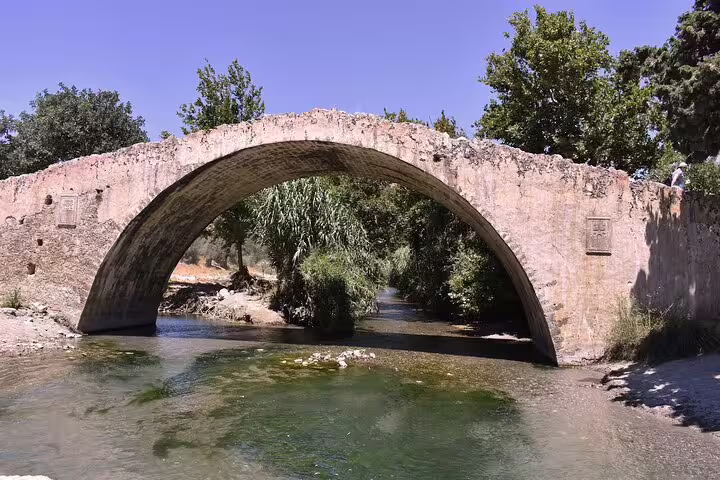 Historic stone arch bridge over a tranquil stream surrounded by lush greenery on Crete's picturesque south coast.