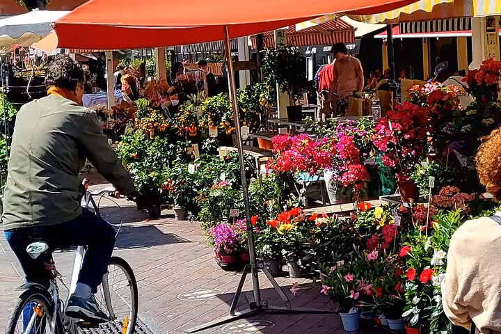 Cyclist exploring a lively flower market in Nice, filled with colorful blooms under bright red umbrellas.