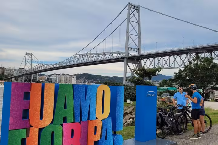 Cyclists near the iconic bridge in Florianopolis, with colorful signage and scenic views enhancing the bike tour experience.