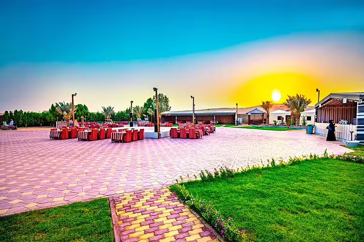 Vibrant sunset over a desert safari campsite in Dubai, featuring BBQ dinner setup with red chairs and live show stage.