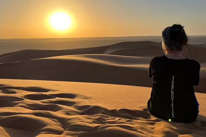 Person sitting on sand dune, gazing at a vibrant desert sunset, ideal for a peaceful camel ride experience.