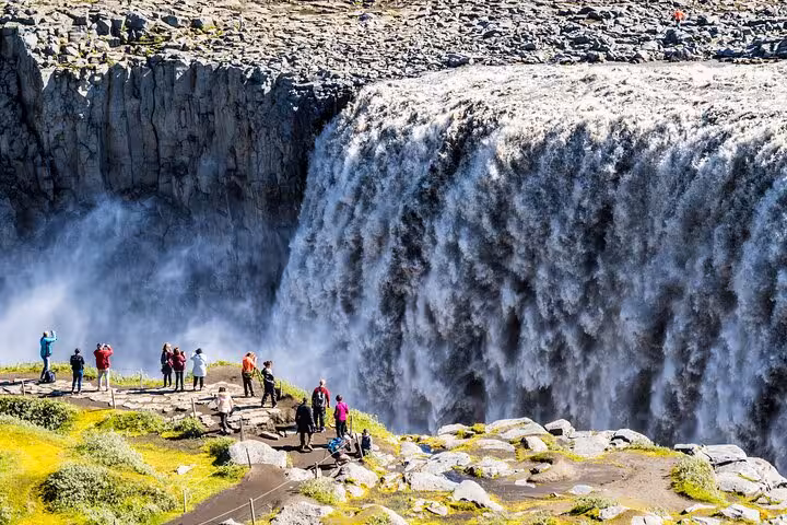 Visitors stand in awe before the powerful, misty cascade of Dettifoss waterfall in Iceland's rugged terrain.