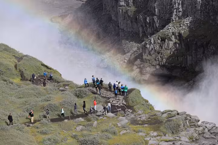 Tourists admire a vibrant rainbow over powerful Dettifoss waterfall in Iceland's dramatic landscape.