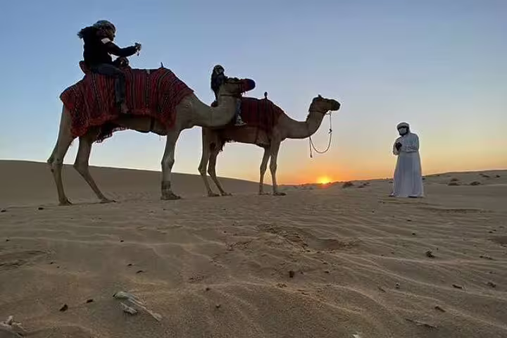 Riders on camels at sunset in the Qatari desert during a private Doha city and desert safari adventure.