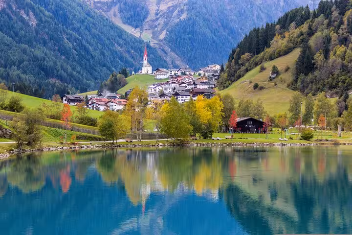 Picturesque village with church, colorful autumn foliage, and serene lake reflecting Dolomites mountains near Venice.