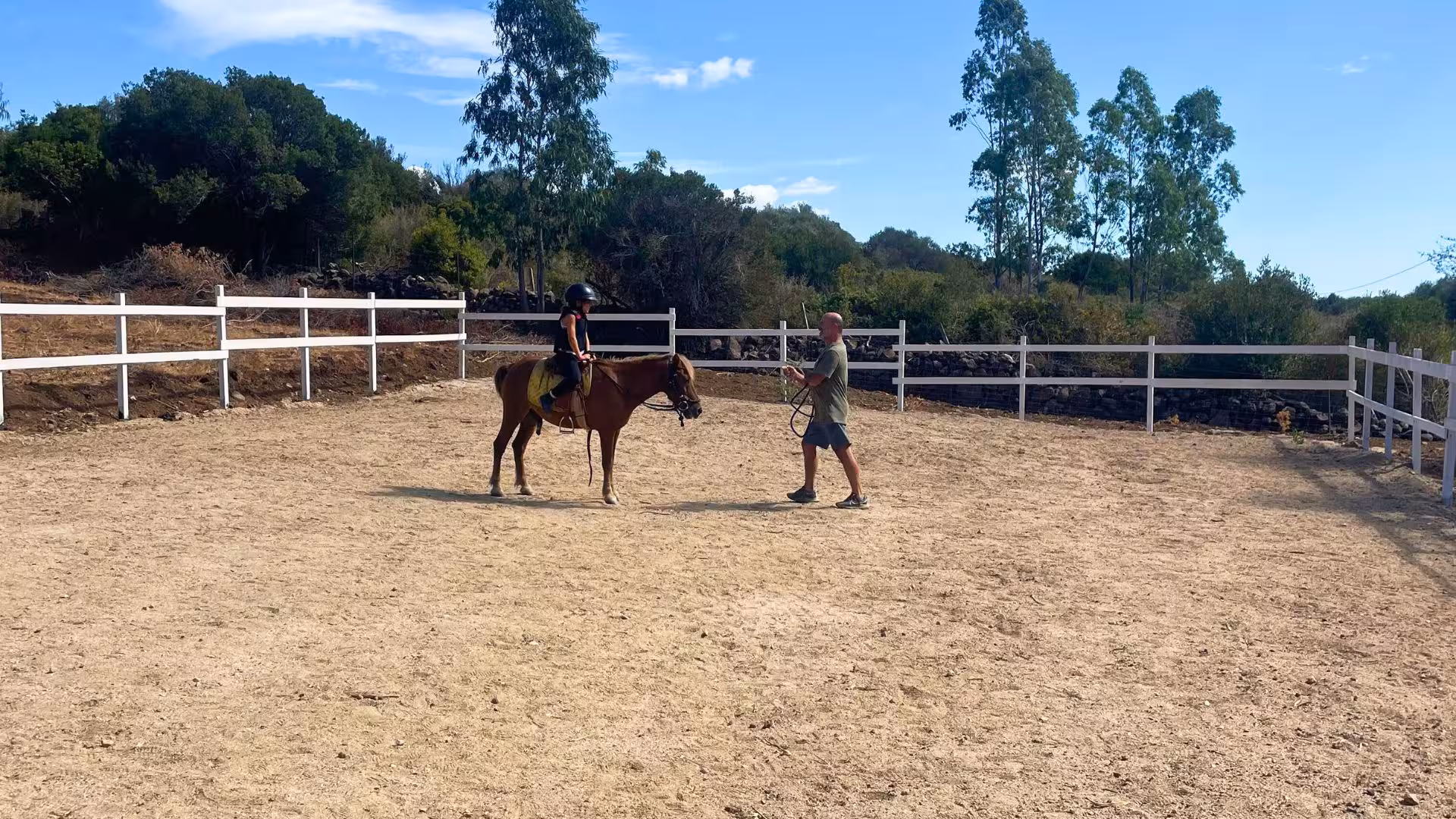 Instructor guides child on a horse in a sunlit arena, ideal for first riding experience in Dorgali.