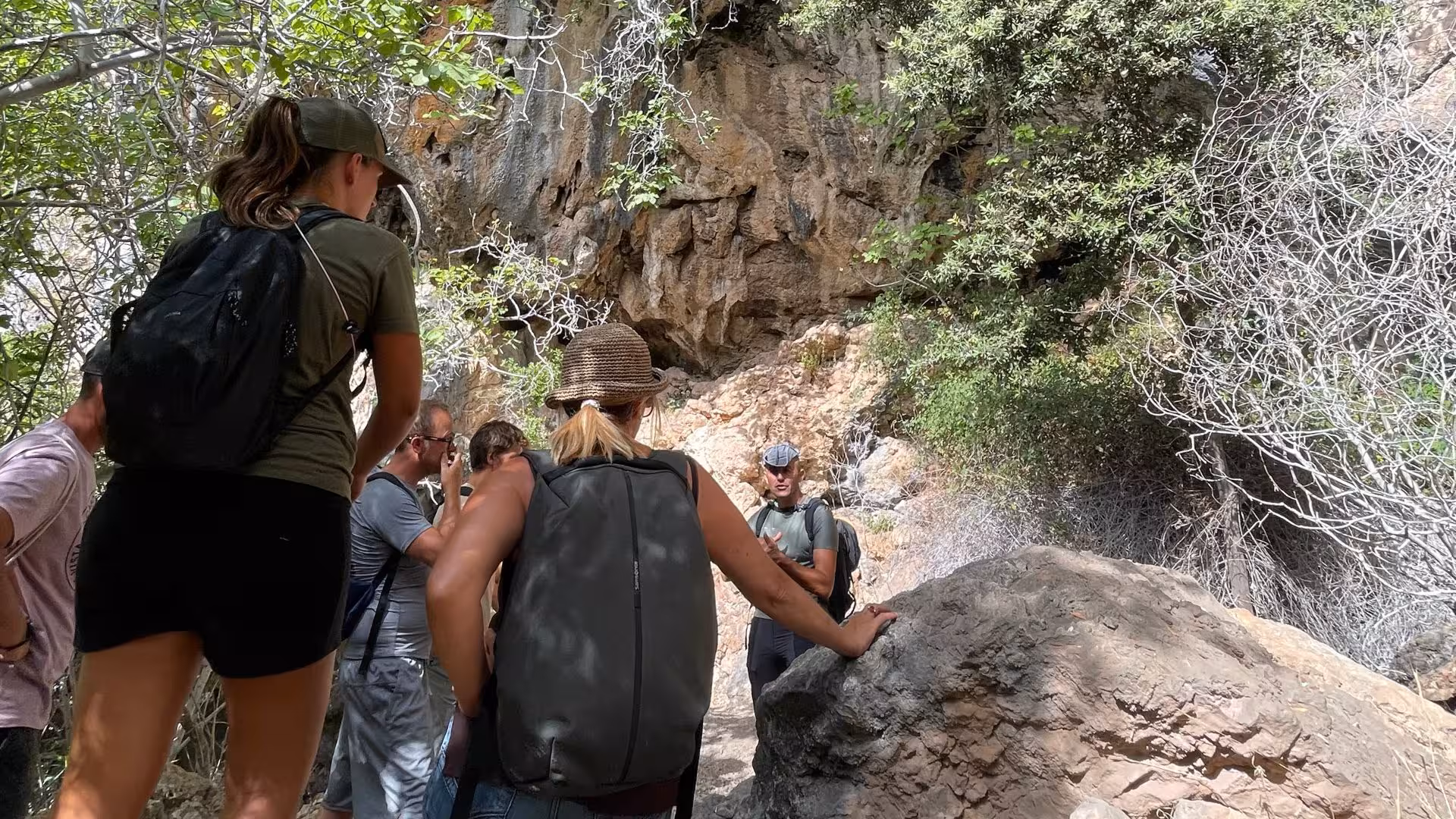 Hiking group listening to a guide in the shaded, rocky terrain of Gorropu Canyon, surrounded by lush vegetation.