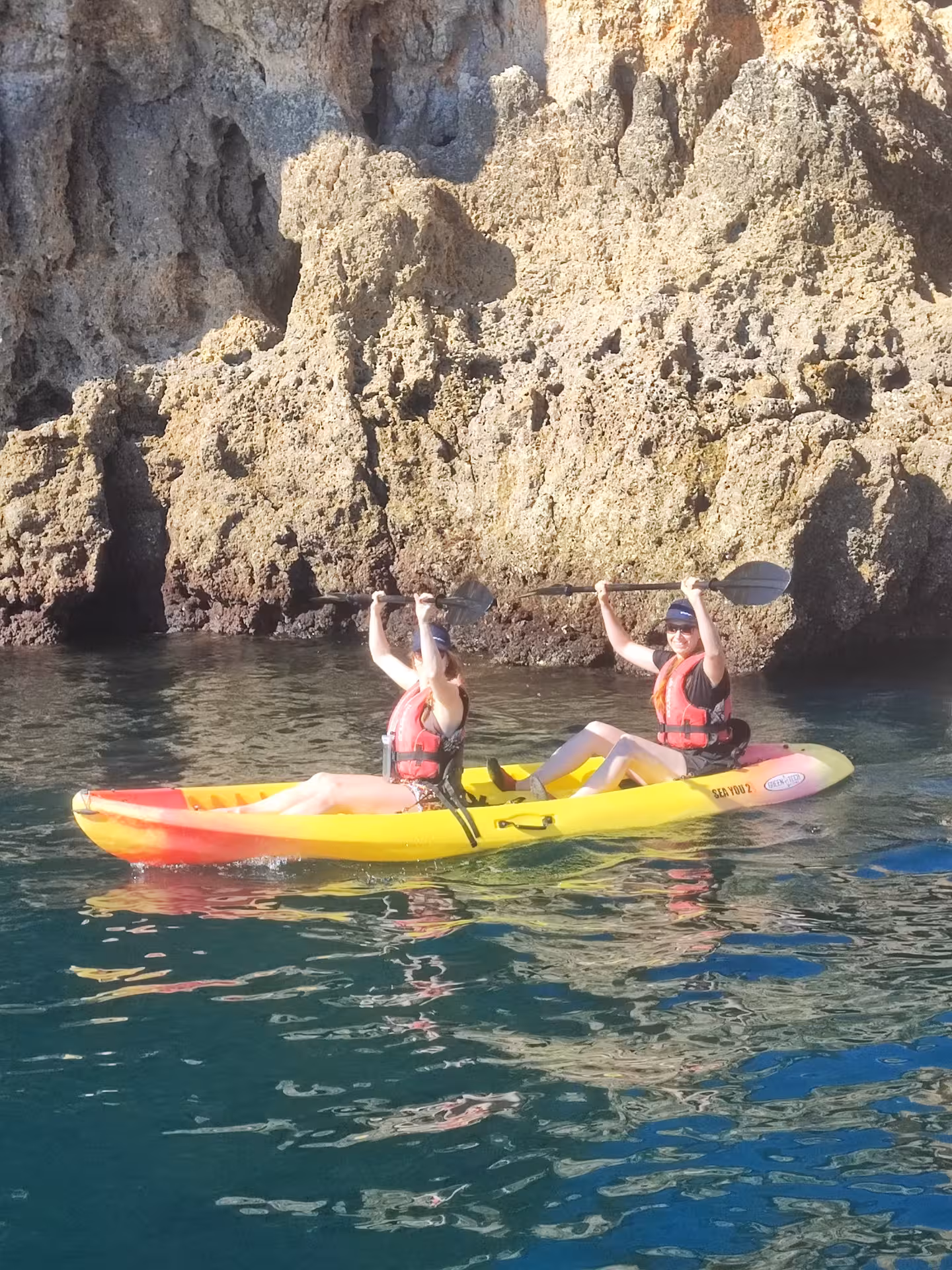 Two women celebrate on a rented double kayak beside rugged Algarve cliffs during a guided Portugal sea kayaking tour