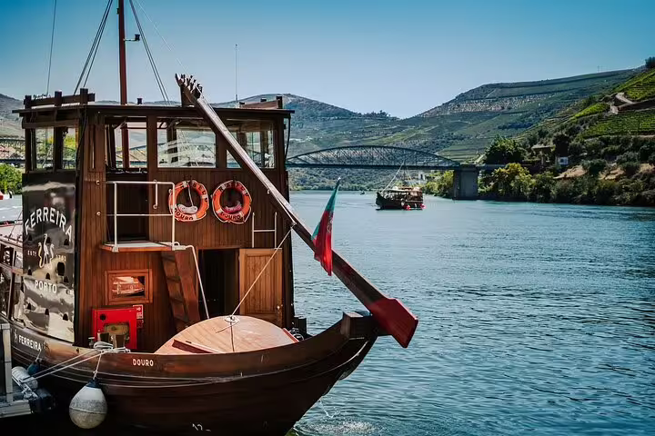 Traditional boat cruising on the Douro River with scenic vineyard hills and a bridge in the background.