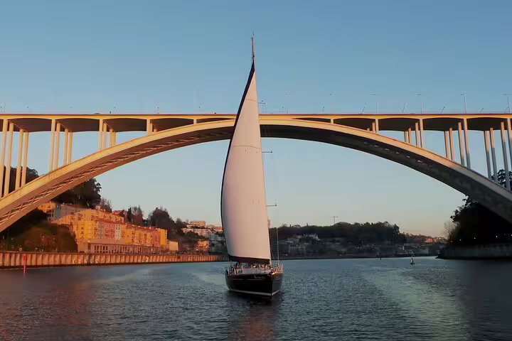 Sailboat gliding under Arrábida Bridge during a picturesque Douro River sunset in Porto, ideal for a scenic sailing experience.