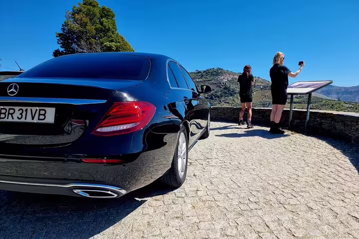 Luxury car parked on a scenic overlook in Douro Valley, Portugal, with tourists capturing vineyard views.