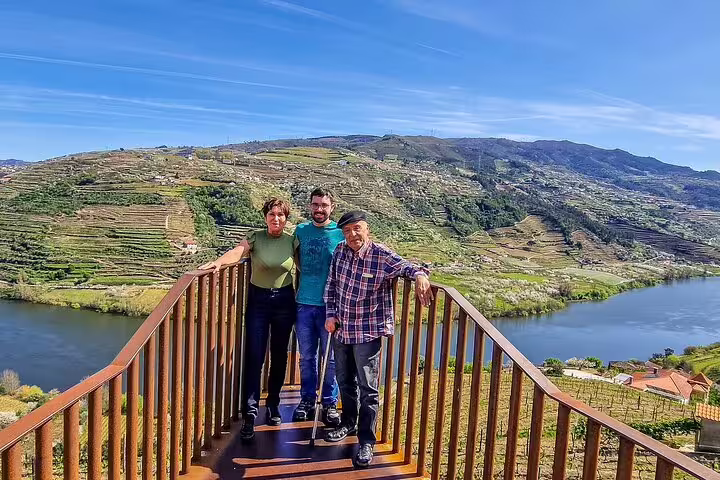 Scenic view of Douro Valley vineyards and river with three tourists enjoying a private tour from Lisbon under a clear blue sky.
